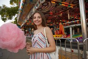 Außenporträt einer charmanten jungen Dame in hellem Kleid mit Trägern, die an warmen Sommertagen über dem Vergnügungspark steht, Zuckerwatte in der Hand hält und mit breitem Lächeln in die Kamera blickt foto