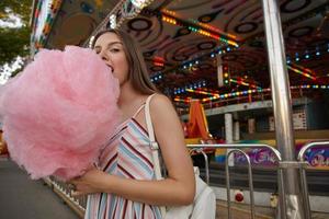 Außenaufnahme einer jungen, langhaarigen, brünetten Frau im Sommerkleid, die über einem Vergnügungspark posiert, Zuckerwatte am Stiel hält und sie beißt, mit geschlossenem Auge in die Kamera schaut foto