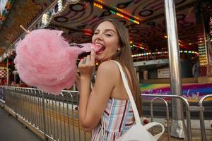 positive junge brünette dame mit langen haaren, die ein kleid mit riemen trägt, über einem vergnügungspark posiert und mit geschlossenem auge und weit geöffnetem mund in die kamera schaut, zuckerwatte hält und sie mit der hand zieht foto