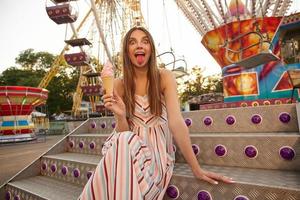 lustiges Outdoor-Foto von hübscher junger Frau im Sommerkleid, die über dem Riesenrad im Vergnügungspark posiert, Eis in der Hand hält und rosa Zunge zeigt foto