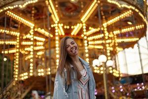 lustiges Outdoor-Foto einer schönen jungen Frau mit langen Haaren, die über Attraktionen im Vergnügungspark posiert, der Kamera zuzwinkert und die Zunge zeigt foto