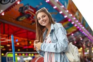 Outdoor-Foto einer positiven jungen hübschen Frau mit langen braunen Haaren, die mit aufrichtigem breitem Lächeln in die Kamera blickt, einen weißen Rucksack trägt, einen trendigen Jeansmantel und ein leichtes Sommerkleid trägt foto