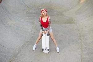 Outdoor-Foto einer hübschen, langhaarigen, tätowierten blonden Frau, die über einem Skatepark posiert, sich an Bord lehnt und die Augen geschlossen hält, legere Kleidung trägt foto
