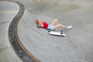 positive junge tätowierte blonde frau im stirnband mit langen haaren, die in lässiger kleidung über dem skatepark liegen, den kopf auf ihren händen halten und fröhlich lächeln foto