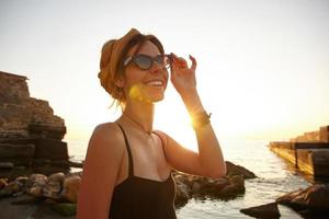 Fröhliche junge hübsche Frau in schwarzem Badeanzug und Stirnband, die ihre Vintage-Sonnenbrille berührt und glücklich beiseite schaut und die Morgendämmerung über dem Meer trifft foto