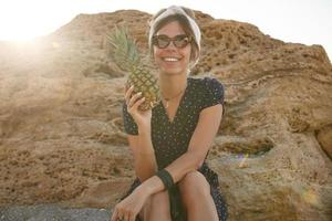 Sonniges Outdoor-Foto einer glücklichen jungen dunkelhaarigen Frau, die mit einem charmanten Lächeln in die Kamera schaut, ein romantisches Sommerkleid und ein Stirnband trägt und frische Ananas in der Hand hält foto