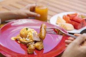 Frauenhand hält eine Gabel und frühstückt. Egg Benedict, Früchte wie Wassermelone, Papaya, Melone, Maracuja, Orangensaft und Kaffee. platziert auf einem grauen Tischset foto