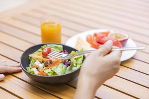 Frauenhand hält eine Gabel und frühstückt. Gemüsesalat, Früchte wie Wassermelone, Papaya, Melone, Maracuja, Orangensaft und Kaffee. platziert auf einem grauen Tischset foto