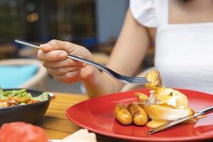 Frauenhand hält eine Gabel und frühstückt. Egg Benedict, Früchte wie Wassermelone, Papaya, Melone, Maracuja, Orangensaft und Kaffee. platziert auf einem grauen Tischset foto