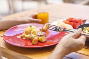 Frauenhand hält eine Gabel und frühstückt. Egg Benedict, Früchte wie Wassermelone, Papaya, Melone, Maracuja, Orangensaft und Kaffee. platziert auf einem grauen Tischset foto