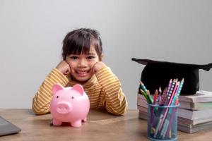 asiatisches kleines mädchen, das eine abschlusskappe mit einem rosa sparschwein trägt, geld spart, die zukunft investiert, foto