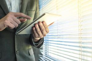 Nahaufnahme eines Geschäftsmannes, der ein Tablet-Gerät verwendet, während er an einem Fenster in einem Büro steht foto