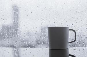 eine tasse kaffee wird morgens mit regentropfen auf dem fenster auf den tisch gestellt. zu hause bleiben und entspannendes konzept. foto