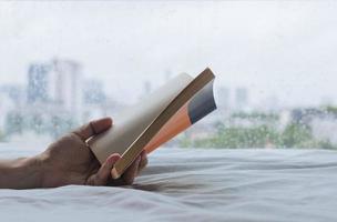 Hand, die ein Buch hält, das an regnerischen Tagen auf dem Bett liest. zu hause bleiben und entspannendes konzept. foto