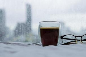 ein glas heißen kaffee mit buch und brille morgens auf dem bett mit regentropfen auf dem fenster. zu hause bleiben und entspannendes konzept. foto