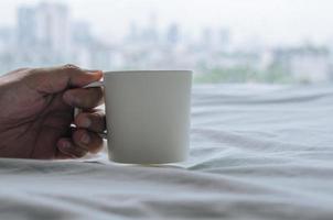 Hand, die an regnerischen Tagen eine Tasse Kaffee auf dem Bett hält. zu hause bleiben und entspannendes konzept. foto