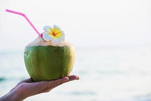 Frische Kokosnuss in Paarhänden mit Plumeria, die am Strand mit Meereswellenhintergrund dekoriert sind - Flitterwochen-Paartourist mit frischem Obst und Meeressand-Sonnenurlaubshintergrundkonzept foto