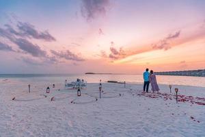 Destination Dining, Flitterwochen, Jubiläum. erstaunlicher romantischer Dinner-Strand auf Holzterrasse mit Kerzen unter Sonnenuntergang. Romantik und Liebe, Luxus-Dinner, exotische Tischdekoration mit Meerblick foto