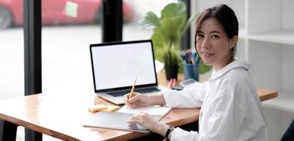 charmante asiatische frau, die im büro arbeitet und einen laptop verwendet, der in die kamera schaut. foto