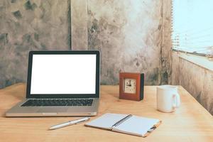 laptop mit weißem leerem bildschirm und tasse kaffee, notizbuch, stift und uhr auf dem hölzernen schreibtisch mit grunge-wandhintergrund. mit Grunge-Wand-Hintergrund. Vintage-Ton foto