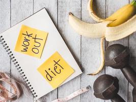 banane mit eisenhanteln, maßband und notizbuch auf dem holztischhintergrund. trainings- und diätkonzept foto