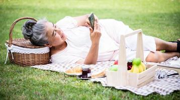 Seniorin, die Smartphone benutzt und nach einem Picknick im Park mit Obst und Essen auf Gras liegt. Gesundheit und Erholung nach der Pensionierung. Seniorenkonzept foto