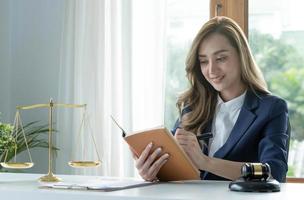 Selbstbewusste und erfolgreiche junge asiatische Rechtsanwältin oder Unternehmensrechtsberaterin, die ein Gesetzbuch liest oder etwas auf ihr Notizbuch an ihrem Schreibtisch schreibt.