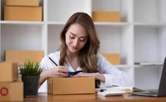 junge schöne glückliche asiatische geschäftsfrau, inhaberin von sme online mit laptop, erhält auftrag vom kunden mit paketverpackung in ihrem start-heimbüro, online-geschäftsverkäufer und lieferung foto