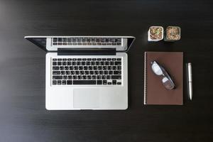 Draufsicht auf Laptop mit Notizbuch, Brille, Stift und Kaktus auf schwarzem Schreibtisch. foto