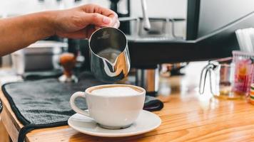 Der professionelle Barista gießt die Milch aus dem Edelstahlbecher in die heiße Kaffeetasse, um im Café einen Cappuccino oder Latte zu machen. Wie man Cappuccino-Kaffee macht foto