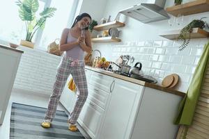 Volle Länge der verspielten Frau mit Schneebesen als Mikrofon beim Kochen in der Küche foto