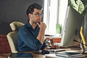 Konzentrierter junger Mann, der Computer benutzt, während er an seinem Arbeitsplatz im Büro sitzt foto