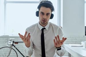 Selbstbewusster junger Mann mit Kopfhörern, der in die Kamera schaut und gestikuliert, während er im Büro sitzt foto