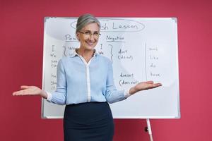 Fröhliche Seniorin, die Englisch in der Nähe des Whiteboards vor rosa Hintergrund unterrichtet foto