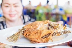 Dame serviert gebratenen Fisch in einem weißen Teller. Das Foto ist auf einen Teil eines Fisches fokussiert.