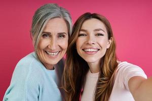 ältere mutter und ihre erwachsene tochter lächeln, während sie selfie vor rosa hintergrund machen foto