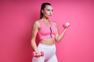 selbstbewusste junge frau in sportkleidung, die mit hanteln vor rosa hintergrund trainiert foto