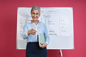 Fröhliche Lehrerin, die die Kamera zeigt, während sie in der Nähe des Whiteboards vor rosafarbenem Hintergrund steht foto