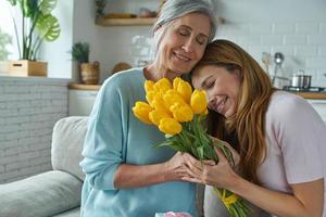 Fröhliche ältere Mutter, die von ihrer Tochter eine Geschenkbox und einen Strauß Tulpen erhält foto