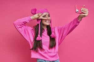 schöne junge frau mit funky hut, die selfie macht und vor rosa hintergrund gestikuliert foto