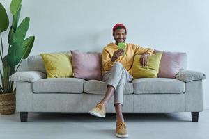 glücklicher afrikanischer mann, der auf sein smartphone schaut, während er auf der couch sitzt foto