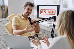 Fröhlicher junger Mann gestikuliert, während er ein Podcast-Interview mit einem Gast im Studio aufnimmt foto