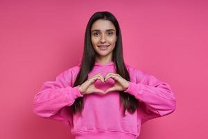 Schöne junge Frau im Kapuzenhemd, die Herzform vor rosa Hintergrund gestikuliert foto