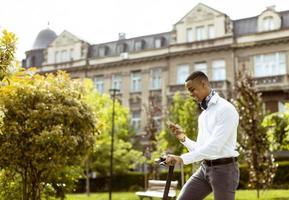 junger Afroamerikaner mit Handy beim Stehen mit Elektroroller auf einer Straße foto