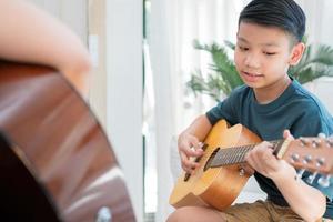 asiatischer junge, der mit vater im wohnzimmer gitarre spielt, um ihm sohn beizubringen, gitarre zu spielen, fühlt sich geschätzt und ermutigt. konzept einer glücklichen familie, lern- und spaßlebensstil, liebe familiäre bindungen foto