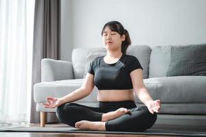 asiatische mollige frau, die auf dem boden im wohnzimmer sitzt, übt yoga-lektion. Frau, die einen Meditationskurs hat. foto