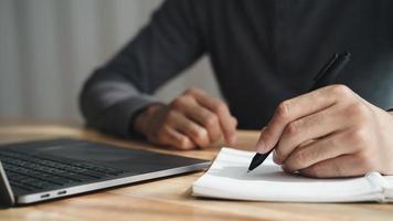 Linkshänder schreibt in einem Notizbuch auf dem Tisch mit Laptop-Computer