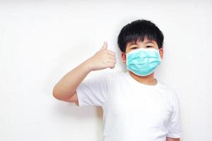 der asiatische junge in einer medizinischen chirurgischen maske, der ein weißes t-shirt trägt und den daumen hoch zeigt. foto