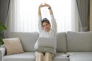 Geschäftsfrau, die ein weißes Hemd trägt und auf dem Sofa sitzt, mit einem Laptop, der ihre Arme über den Kopf hebt, um Schmerzen zu lindern, nachdem sie lange bei der Arbeit gesessen hat. foto