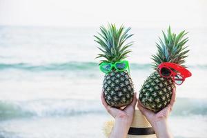 schönes paar frische ananas, die jungen- und mädchenbrillen in touristische hände mit seewellenhintergrund legen - fröhlicher spaß mit gesundem urlaubskonzept foto